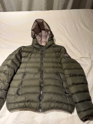 Olivgrön pufferjacka från Colmar - Säljer en olivgrön pufferjacka från Colmar Originals med huva och två dragkedjeförsedda fickor. Jackan har quiltad design och beige insida, perfekt för kalla dagar. Klassisk logga på armen och smidig dragkedja framtill.