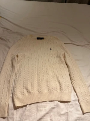 Vit kabelstickad tröja Polo Ralph Lauren - Kabelstickad vit tröja från Polo Ralph Lauren med klassisk rund hals och broderad logga på bröstet. Tröjan har ribbade muddar och är långärmad