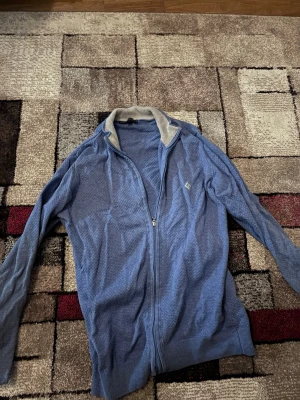 Blå kofta från Sir Raymond Tailor - Blå kofta med dragkedja från Sir Raymond Tailor i storlek S. Tröjan har ett diskret mönster i tyget och ribbad krage i beige. Långärmad och med liten logga på bröstet. Perfekt för dig som gillar stilrena och bekväma plagg. Lite urtvättad och små fläckar som går bort i tvätten.