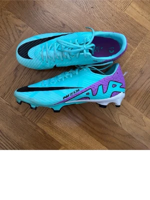 Nike Mercurial Air Zoom fotbollsskor - Säljer ett par Nike Mercurial Air Zoom fotbollsskor i en riktigt fräsch turkos färg med lila detaljer och svart swoosh. Skorna har snörning, låg profil och mönstrad sula för gräsplan. Materialet är syntet och designen är riktigt sportig och modern. Skorna 