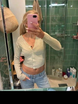 Beige croppad kofta med knappar - Säljer en beige croppad kofta med v-ringning och långa ärmar. Koftan har ribbad struktur och stängs med vita knappar framtill. Perfekt att styla med jeans eller kjol för en chill look.