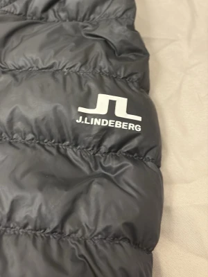 Svart dunjacka från J.Lindeberg - Säljer en svart dunjacka från J.Lindeberg med huva och dragkedja. Jackan är quiltad och har två sidofickor med dragkedja. Perfekt för kalla dagar och riktigt lätt att bära. Klassisk och stilren design med logga på bröstet.