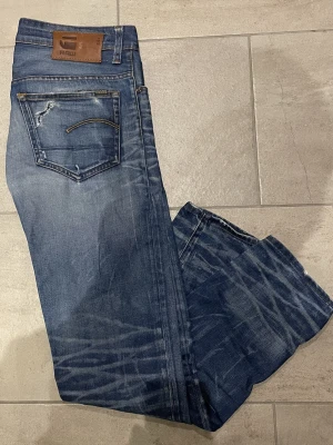 Blå G-Star RAW jeans, storlek 31/32 - Säljer ett par blå jeans från G-Star RAW i storlek 31/32. Jeansen har slitna detaljer, klassisk femficksmodell och coola tvättade effekter på benen. Materialet är robust denim och passformen är rak med normal midja. Perfekta för en avslappnad streetstil.