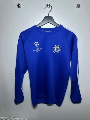 Chelsea Adidas blå träningströja - Chelsea FC långärmad träningströja från Adidas med Champions League-logga och ClimaCool-tyg. Tröjan är klarblå med vita detaljer och ränder längs ärmarna samt stort 'CHELSEA' tryckt på ryggen. Perfekt för dig som älskar fotboll och vill ha en ikonisk look.
