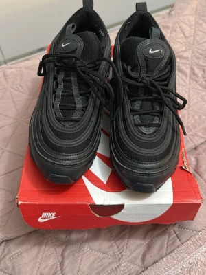 Nike Air Max 97 Triple Black sneakers - Säljer ett par Nike Air Max 97 i färgen Triple Black. Skorna har en ikonisk vågig design, synlig Air-enhet i sulan och reflekterande detaljer. Ovandelen är i mesh och syntet med svarta snören och små vita Swoosh-loggor på sidan och tungan.