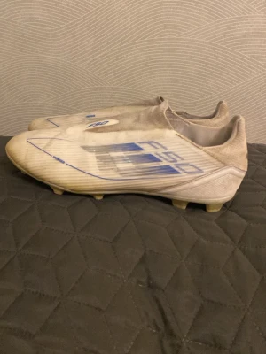 Adidas F50 vita fotbollsskor - Säljer ett par Adidas F50 fotbollsskor i vitt med blå detaljer. Skorna har en strömlinjeformad design utan snörning och är tillverkade i syntetmaterial för lätt vikt och bra bollkontroll. Perfekta för dig som vill ha snabba fötter på planen.