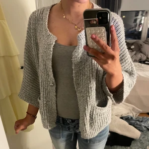 Grå stickad kofta med fickor - Mysig grå stickad kofta med grov struktur och trekvartsärmar. Koftan har två praktiska fickor framtill och en öppen design utan knappar. Perfekt att slänga på sig över ett linne eller t-shirt för en chill look.