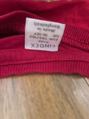 Röd jul-tshirt sopran hundmotiv Lindex - Röd t-shirt från Lindex med ett stort tryck av en söt hund i tomteluva och texten 'Merry Xmas' i guld. T-shirten har korta ärmar och är gjord i mjuk bomull, perfekt för julens alla tillfällen.