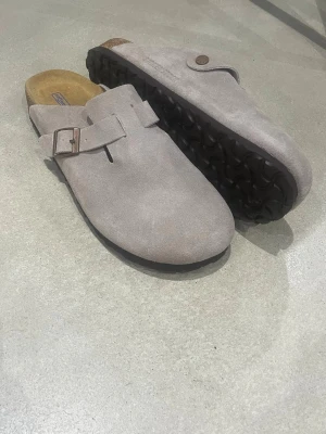 Grå mocka Birkenstock Boston - Säljer ett par Birkenstock Boston i ljusgrå mocka med justerbart spänne i metall. Skorna har en klassisk korksula och svart räfflad yttersula. Perfekta för chill dagar och riktigt sköna att glida runt i.