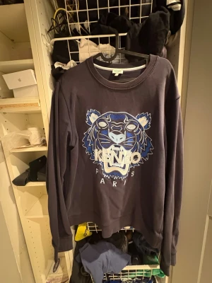 Mörkblå Kenzo sweatshirt med tiger - Säljer en mörkblå sweatshirt från Kenzo i storlek L. Tröjan har det ikoniska broderade tigermotivet i blått, vitt och beige på bröstet samt ribbade muddar vid hals, ärmslut och nederkant. Perfekt statement-plagg för dig som gillar streetwear.