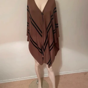 Brun poncho med svarta ränder - Säljer en brun poncho med svarta diagonala ränder och v-ringad hals. Ponchon har en lös och flowy passform och stängs framtill med en knapp. Perfekt för lager på lager under höst och vår.