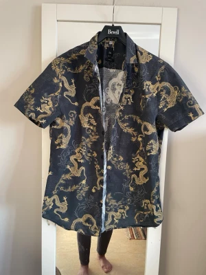 Herr skjorta med guldiga drakar River Island - Säljer en kortärmad skjorta från River Island i slim fit med ett fett guldigt drakmönster över hela. Skjortan har knappar framtill och klassisk krage. Perfekt för dig som vill sticka ut med en unik stil.
