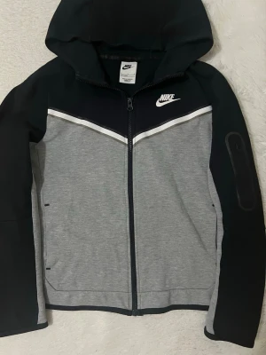 Nike sportig vindjacka med huva (Nike tech fleece) - Nike vindjacka i svart och grått med vit detalj över bröstet. Jackan har huva, hel dragkedja och praktiska fickor. Snygg sportig look med logga på bröstet och en extra ficka på ärmen. Perfekt för dig som gillar streetwear och sportig stil. (Nike tech) size 147-158cm. Blev för liten för mig  så jag vill sälja den istället för att kasta.