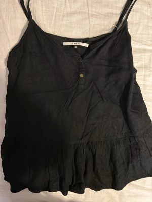Svart linnetopp med volang från ONLY - Säljer en svart linnetopp från ONLY i storlek 38. Toppen har smala axelband, knappar framtill och en bred volang längst ner som ger en söt touch. Perfekt till sommaren och lätt att matcha med olika stilar.