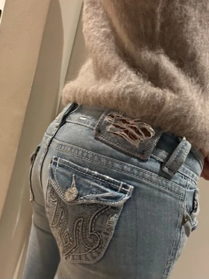 Ljusblå bootcut jeans från MEK Denim - Säljer ett par ljusblå jeans från MEK Denim med snygga broderade detaljer på bakfickorna och slitningar på linningen. Modellen har låg midja och bootcut-ben, vilket ger en retro och avslappnad vibe. Klassisk femficksdesign och coola kontrastsömmar.