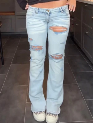 Ljusblå bootcut jeans med slitningar - Säljer ett par ljusblå jeans med bootcut passform och stora slitningar på låren och knäna. Jeansen har låg midja och klassisk femficksdesign. Perfekta för dig som gillar en avslappnad och trendig look.