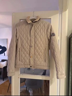 Beige quiltad jacka från Henri Lloyd - Säljer en beige quiltad jacka från Henri Lloyd på grund av flytt.  Snyggt rutmönster och mjuk manchesterkrage. Jackan har dragkedja och knappar framtill, samt två fickor. Perfekt för dig som vill ha en stilren och klassisk look.
