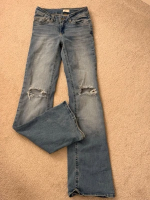 Blå bootcut jeans med slitna knän - Säljer ett par ljusblå bootcut jeans med slitningar över knäna och snygga detaljer i form av guldfärgade nitar på bakfickorna. Dom är lågmidjade och knappt använda, säljer pga av att dom är för långa💖