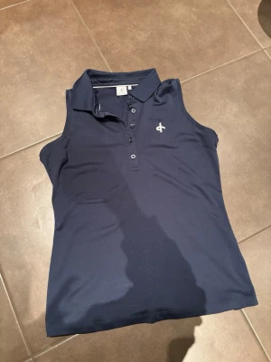 Marinblå ärmlös pikétopp Cross - Säljer en marinblå ärmlös pikétopp från Cross Sportswear. Toppen har klassisk krage, knappar framtill och ett diskret broderat logomärke på bröstet. Tillverkad i ett mjukt och stretchigt syntetmaterial som andas, perfekt för sportiga dagar.