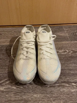 Nike vita fotbollsskor Air Zoom - Säljer ett par vita Nike Air Zoom fotbollsskor med glansig finish och svarta detaljer. Skorna har snörning, rund tå och är tillverkade i syntetmaterial med stickad ovandel. Perfekta för dig som vill sticka ut på planen med stilrena och moderna skor.