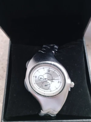 Y2K / Organic Future Style - Säljer en klocka med den perfekta "Y2K"-looken. Designen är organisk och futuristisk med borstad silverfinish.                                                                       Stil: Tänk "liquid metal" eller 00-tals futurism. Urtavlan har en sportig "racing"-look med subtiles-dial detaljer. Skick: Helt ny och oanvänd, skyddsplasten sitter kvar på länken. Detaljer: Fin, borstad metallfinish (inte blank/kromad) vilket ger en lyxigare känsla. För dig som gillar den unika 2000-tals estetiken