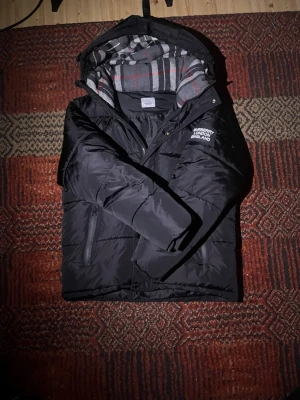 Svart pufferjacka från Burberry - Säljer en svart pufferjacka från Burberry med avtagbar huva och klassiskt rutigt foder i grått, vitt och rött. Jackan har dragkedja, tryckknappar och en tydlig Burberry London England-logga på bröstet. Perfekt för kalla dagar och riktigt snygg streetstil. Vissa va du har att erbjuda så kanske vi kan diskutera fram ett pris där både jag och du är nöjda 