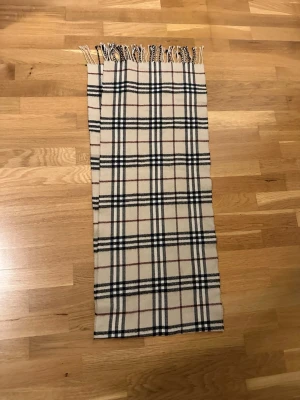 Beige rutig halsduk med fransar - Klassisk beige halsduk med rutigt mönster i svart, vitt och rött. Halsduken har fransar i ändarna och är tillverkad i ett mjukt material som känns skönt mot huden. Perfekt accessoar för att lyfta din outfit.