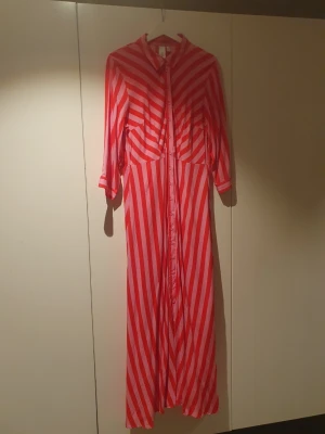 Röd- och rosarandig långklänning Y.A.S - Säljer en oanvänd långklänning från Y.A.S i storlek XL. Klänningen har röd- och rosarandigt mönster, knappar hela vägen framtill, krage och trekvartsärm. Materialet känns lätt och flowy, perfekt för en färgstark look.