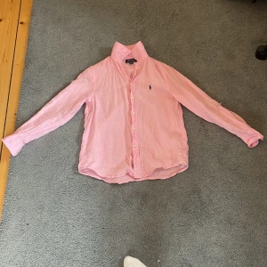 Rosa skjorta från Polo Ralph Lauren - Snygg rosa skjorta från Polo Ralph Lauren med klassisk krage och knappar framtill. Skjortan har lång ärm och är tillverkad i mjukt bomullstyg. På bröstet finns den ikoniska blå broderade Polo-loggan. Perfekt för dig som gillar stilrena och färgglada plagg. Ny pris 1200