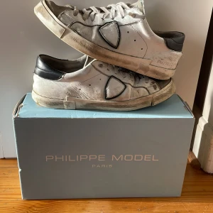 Philippe Model vita sneakers med svarta detaljer - Snygga vita sneakers från Philippe Model med svarta detaljer på häl och sidor. Skorna har snörning, rund tå och platt sula. Tillverkade i skinn med mockadetaljer på tån. Perfekta för dig som gillar en klassisk men trendig look.