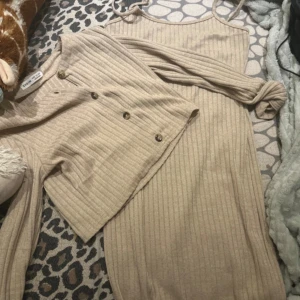 Beige ribbad långklänning från Shein - Säljer en beige ribbad långklänning från Shein med smala axelband och matchande cardigan med knappar. Klänningen har en mjuk stickad känsla och är perfekt för dig som gillar en chill och stilren look.