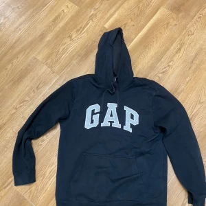 Svart hoodie från GAP med logga - Svart hoodie från GAP med stor grå logga framtill. Tröjan har huva med snörning, magficka och långa ärmar. Perfekt för chill dagar och enkel att matcha med jeans eller joggers. Mjuk bomullskänsla och klassisk passform.