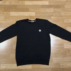 Svart sweatshirt från Moncler - Svart sweatshirt från Moncler i storlek M med rund hals och ribbade muddar. Tröjan har ett broderat Moncler-logo på bröstet och en diskret röd-vit-blå detalj på insidan av kragen. Perfekt för dig som gillar stilrena och exklusiva plagg.
