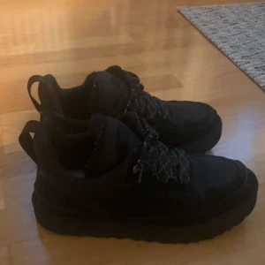 Ugg lowmel storlek 44  -  Använda fåtal gånger förra vintern. Svarta sneakers från UGG med grov platt sula och snörning med mönstrade svarta och vita skosnören. Insidan är fodrad med mjuk syntetpäls för extra komfort. Ovandelen är i mocka och textil, perfekt för höst och vinter.