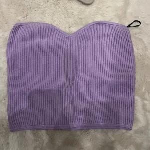 Ljuslila ribbad tubtopp - Säljer en ljuslila ribbad tubtopp med bandeauform. Toppen är ärmlös och har en mjuk, stretchig passform som sitter snyggt mot kroppen. Perfekt att styla med jeans eller kjol för en trendig look.