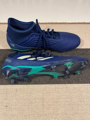Adidas Predator 18.3 blå fotbollsskor - Säljer ett par Adidas Predator 18.3 fotbollsskor i marinblått med turkosa detaljer och vita ränder. Skorna har snörning, platt sula med dobbar och en stickad, strumpliknande krage för extra stöd. Tillverkade i syntetmaterial och designade för gräsplaner.