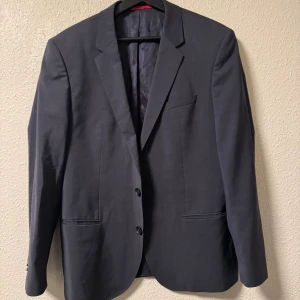 Svart kavaj från Hugo Boss - Stilren svart kavaj från Hugo Boss med klassisk passform. Kavajen har två knappar framtill, smala slag och innerfoder i glansigt material. Perfekt för dig som vill ha en tidlös och snygg look. Märke i rött på insidan.