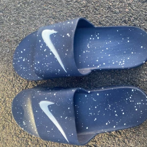 Nike Vapormax sandaler blå/vita - Nike Vapormax sandaler i mörkblå färg med vita stänk och stor vit Swoosh-logga. Unik sula med Vapormax-detaljer och text på sidan. Tillverkade i syntetmaterial, platt sula och öppen tå. Perfekt för chill dagar och sommarhäng.