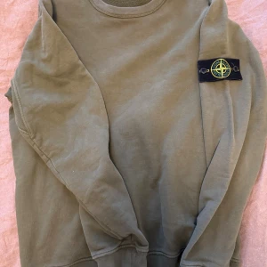 Olivgrön Stone Island sweatshirt - Säljer en olivgrön sweatshirt från Stone Island med den klassiska patchen på vänster ärm. Tröjan har rund hals och ribbade muddar vid ärmslut och nederkant. Materialet är mjuk bomull som är skön mot huden. .