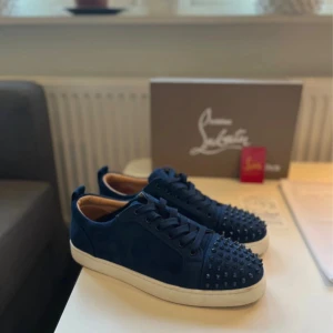 Mörkblå Christian Louboutin - Säljer ett par mörkblå sneakers från Christian Louboutin med coola nitar på tån och vit platt sula. Skorna har snörning och är gjorda i mocka med lyxig känsla. Insidan är beige och de har en rund tå. Perfekt för dig som vill sticka ut med designerstil.ops en nit är limad
