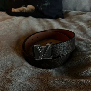 Louis Vuitton monogram bälte brun Helt nytt - Snyggt Louis Vuitton bälte (helt nytt) i brunt skinn med klassiskt monogrammönster och silverfärgat LV-spänne. Bältet har en stilren design med tydliga LV-loggor och detaljer i beige på insidan. Perfekt accessoar för att lyfta din outfit.