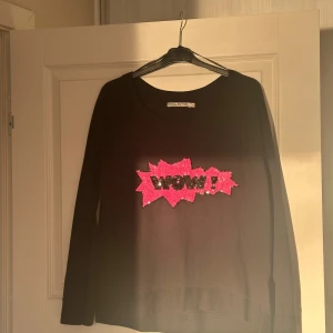 Svart tröja med WOW-paljetter Soul River - Svart långärmad tröja från Soul River i 100% bomull. Framtill finns en stor applikation med rosa och svarta paljetter som bildar texten 'WOW!'. Tröjan har rund halsringning och en avslappnad passform. Perfekt för dig som gillar statement-detaljer.