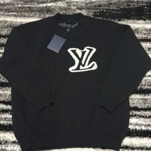 Svart stickad tröja Louis Vuitton - Svart stickad tröja från Louis Vuitton med stort vitt LV-logo på bröstet och ryggen. Klassisk rund halsringning och ribbade muddar vid ärmslut och nederkant. Perfekt för dig som vill ha en lyxig och stilren look.