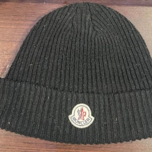Svart ribbad Moncler mössa - Svart ribbstickad mössa från Moncler med klassisk logotyp framtill. Mössan är tillverkad i mjuk ull och har en uppvikt kant för extra stilpoäng. Perfekt för dig som vill ha en clean och lyxig look under kyliga dagar.