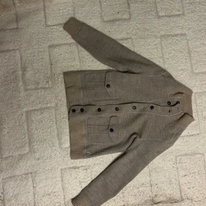 Beige kofta från Eleventy i ull - Snygg beige kofta från Eleventy med ribbade muddar och krage. Tröjan har knäppning framtill med svarta knappar och två stora fickor på bröstet. Tillverkad i mjuk ull, perfekt för lager-på-lager under kyliga dagar. Kostar runt 7000 till 10000 ny. Den ser grå ut på bilderna men är mer beige i verkligeheten! Passar runt 160- lite kortare än 170