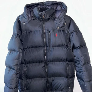 Svart pufferjacka från Polo Ralph Lauren - Svart pufferjacka från Polo Ralph Lauren med röd broderad logga på bröstet. Jackan har huva, flera dragkedjor och en quiltad design som ger en schysst volym. Materialet är polyester och dun, perfekt för kalla dagar. Snygg och klassisk look med normal passform.
