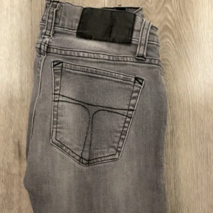 Grå jeans från Tiger of Sweden - Snygga grå jeans från Tiger of Sweden med klassisk femficksdesign och svarta detaljer. Jeansen är tillverkade i 98% bomull och 2% elastan för en bekväm passform. Stilen är rak och tidlös, perfekt för dig som gillar en clean look.