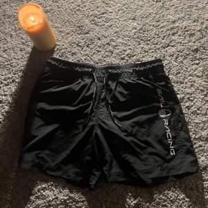 Svarta Sail Racing shorts med snörning - Svarta shorts från Sail Racing med vit logga längs benet och snörning i midjan. De har en loose passform och är gjorda i lätt polyester, perfekta för sommarens alla äventyr. Snygg sportig look med elastisk midja och coola detaljer på snörena.