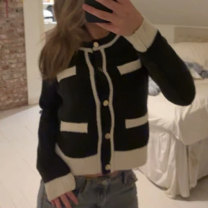 Svartvit kofta från Vero Moda - Snygg svart kofta med vita detaljer från Vero Moda. Koftan har rund hals, dekorativa fickor och guldknappar framtill. Ribbstickade muddar och kontrastkanter ger en cool och stilren look. Perfekt att slänga över en t-shirt eller linne.