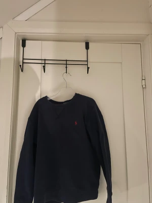 Mörkblå sweatshirt från Polo Ralph Lauren str 10-12y - Klassisk mörkblå sweatshirt från Polo Ralph Lauren med rund halsringning och röd broderad logga på bröstet. Tröjan har långa ärmar och en avslappnad passform, perfekt för en chill och stilren look.
